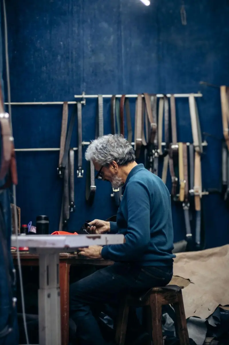 Leather craftsman working by hand in a workshop surrounded by leather straps, representing European artisan slow production