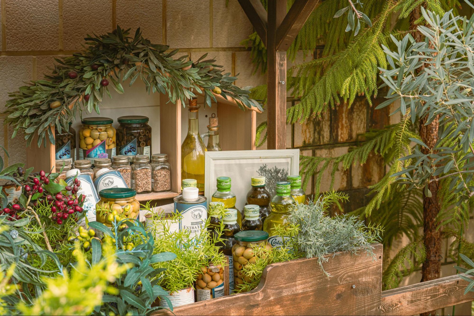 European artisan market display with olive oil bottles, olives in glass jars, and fresh herbs arranged on a rustic wooden shelf